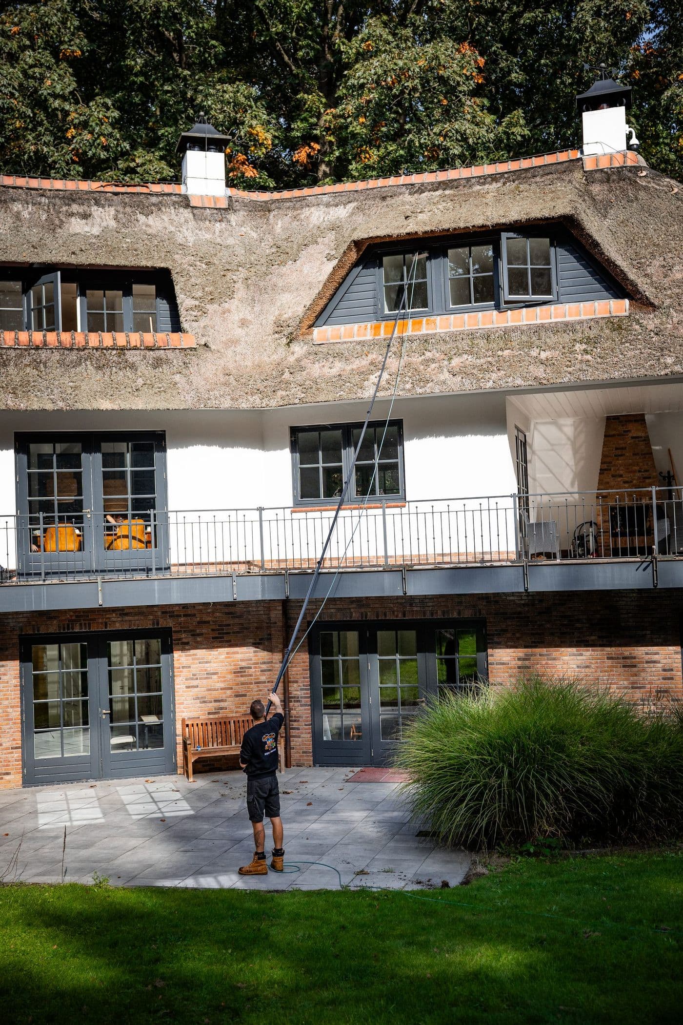 Medewerker reinigt met een telescoopsteel de ramen van een villa met rieten dak